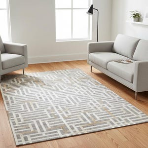 Puede incluir: Una moderna sala de estar con un sofá y un sillón grises claros con patas de metal. Una alfombra estampada en tonos grises, blancos y beige cubre el suelo. Una lámpara negra y una estantería blanca con decoración completan la escena.