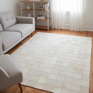 Puede incluir: Una alfombra rectangular grande con un diseño de patchwork en tonos blancos y crema. La alfombra está colocada sobre un suelo de madera, con un sofá y una silla grises en el fondo. La textura y el patrón de la alfombra añaden un toque de elegancia a la habitación.