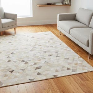 May include: A rectangular area rug with a geometric pattern of triangles in shades of beige, cream, and gray. The rug is placed on a hardwood floor, with a gray sofa and armchair in the background.