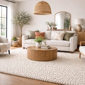May include: A bright, airy living room with a white sofa, two armchairs, and a round wooden coffee table. The room features a large, textured rug, a woven pendant light, and decorative plants. The color palette is neutral with pops of green and orange.