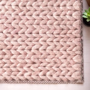 May include: A close-up of a soft, chunky knit rug in a light pink colour. The rug has a braided pattern, with each stitch clearly visible. The texture appears plush and cosy, suitable for a living room or bedroom.