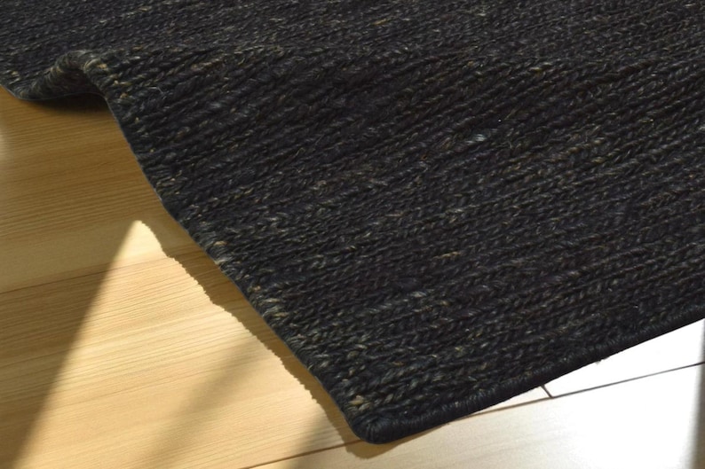 May include: Close-up of a dark charcoal-colored rug with a woven texture. The rug's edge is visible, showing a tightly bound border. The rug is placed on a light-colored wooden floor, with a shadow cast across the surface.