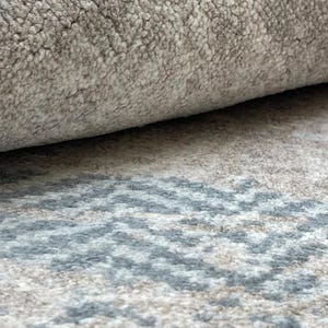 May include: Close-up of a rolled-up rug with a textured surface. The rug features a neutral color palette with shades of gray and beige. A geometric pattern in blue and gray is visible on the unrolled portion.