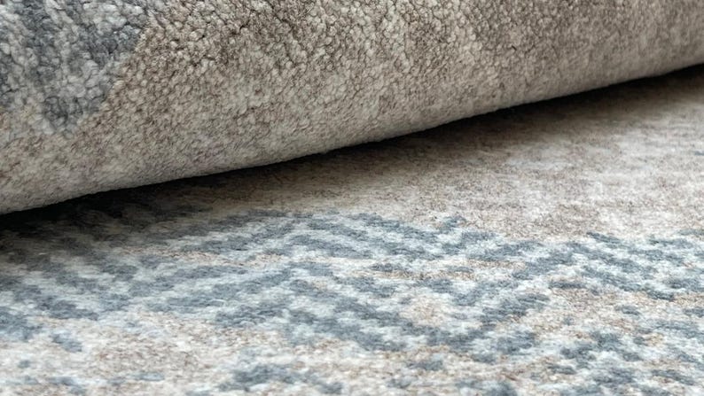May include: Close-up of a rolled-up rug with a textured surface. The rug features a neutral color palette with shades of gray and beige. A geometric pattern in blue and gray is visible on the unrolled portion.