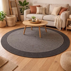 Puede incluir: Una alfombra redonda gris oscuro con círculos concéntricos en un patrón gris claro. La alfombra tiene un borde gris oscuro y está colocada sobre un suelo de madera. Una mesa de centro de madera está sobre la alfombra.