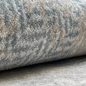 May include: Close-up of a rolled-up area rug. The rug features a textured pattern in shades of gray, beige, and blue. The edges of the rug are visible, showcasing the thickness and texture of the material. The rug appears to be made of a soft, durable material.