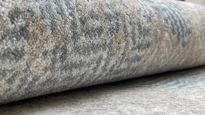 May include: Close-up of a rolled-up area rug. The rug features a textured pattern in shades of gray, beige, and blue. The edges of the rug are visible, showcasing the thickness and texture of the material. The rug appears to be made of a soft, durable material.