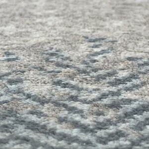 May include: Close-up of a textured rug with a subtle pattern. The rug features a blend of gray, beige, and light blue hues, creating a soft, neutral aesthetic. The design has a subtle, repeating pattern.