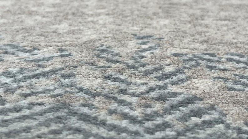 May include: Close-up of a textured rug with a subtle pattern. The rug features a blend of gray, beige, and light blue hues, creating a soft, neutral aesthetic. The design has a subtle, repeating pattern.