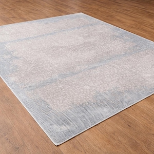 May include: A rectangular area rug with a light gray center and a blue border. The border features a herringbone pattern. The rug is placed on a wooden floor, creating a contrast in textures and colors.