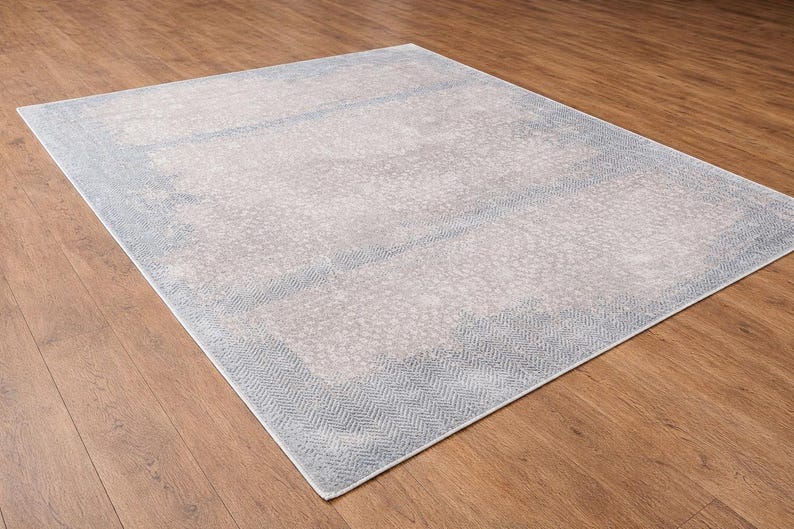 May include: A rectangular area rug with a light gray center and a blue border. The border features a herringbone pattern. The rug is placed on a wooden floor, creating a contrast in textures and colors.