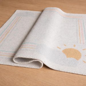 May include: A white bath mat with a rolled edge, featuring a sun graphic in the corner. The mat has a rectangular shape with pastel-coloured stripes along the border, adding a touch of colour to the otherwise neutral design.