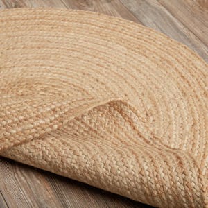 May include: A natural-toned, round jute rug with a woven texture. The rug is partially rolled up, revealing the underside and the intricate weave pattern. The rug is made of a light brown material and is suitable for home decor.
