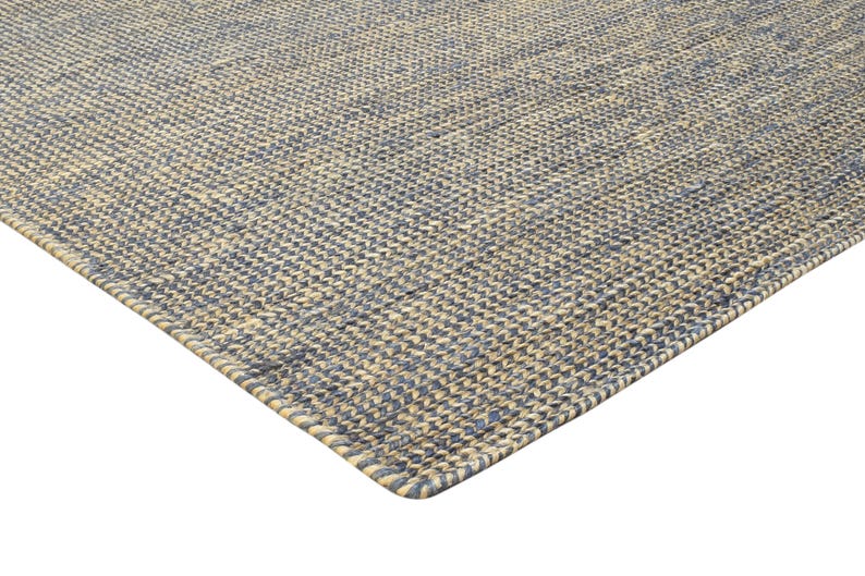 Puede incluir: Vista en esquina de una alfombra tejida con una superficie texturizada. La alfombra presenta un patr&oacute;n de hilos azules y beige alternados, creando un dise&ntilde;o geom&eacute;trico sutil. Los bordes est&aacute;n cuidadosamente acabados.