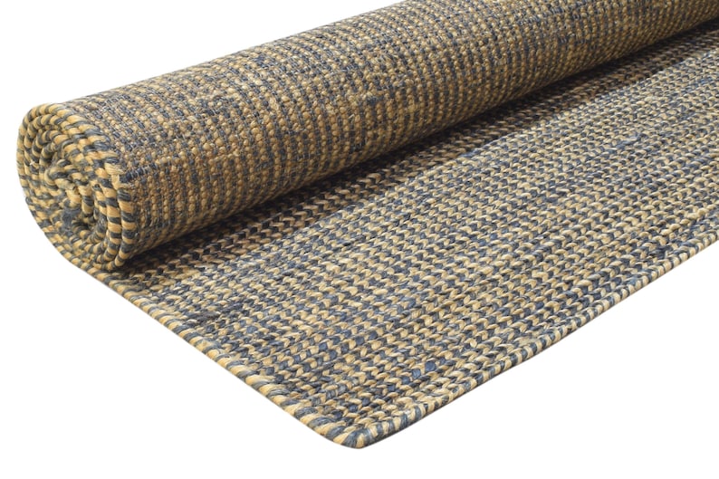 Puede incluir: Una alfombra enrollada con un patr&oacute;n tejido en tonos beige y azul marino. La alfombra est&aacute; hecha de una fibra natural y tiene un aspecto texturizado. El borde de la alfombra es visible, mostrando el grosor y el tejido.