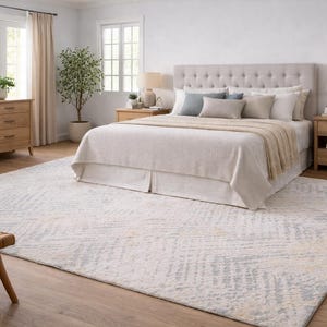 May include: A bedroom with a neutral color palette, featuring a large bed with a tufted headboard, a wooden dresser, and a patterned rug. The room is well-lit with natural light from the windows.