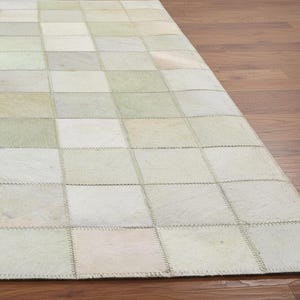 May include: A rectangular rug with a patchwork design. The rug is composed of numerous squares in shades of white, cream, and pale green. The squares are stitched together, creating a textured surface. The rug is placed on a wooden floor.