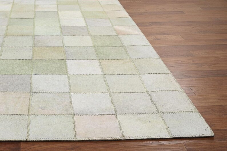 May include: A rectangular rug with a patchwork design. The rug is composed of numerous squares in shades of white, cream, and pale green. The squares are stitched together, creating a textured surface. The rug is placed on a wooden floor.