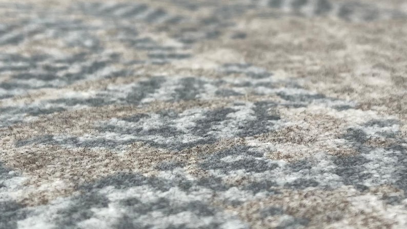 May include: Close-up of a textured rug with a repeating pattern. The rug features a mix of gray, white, and brown tones, creating a neutral and versatile design. The fibers appear soft and plush, suggesting comfort.