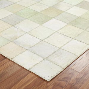 May include: A patchwork rug with a geometric pattern of squares in shades of cream, beige, and pale green. The rug is made of stitched-together pieces and rests on a wooden floor.