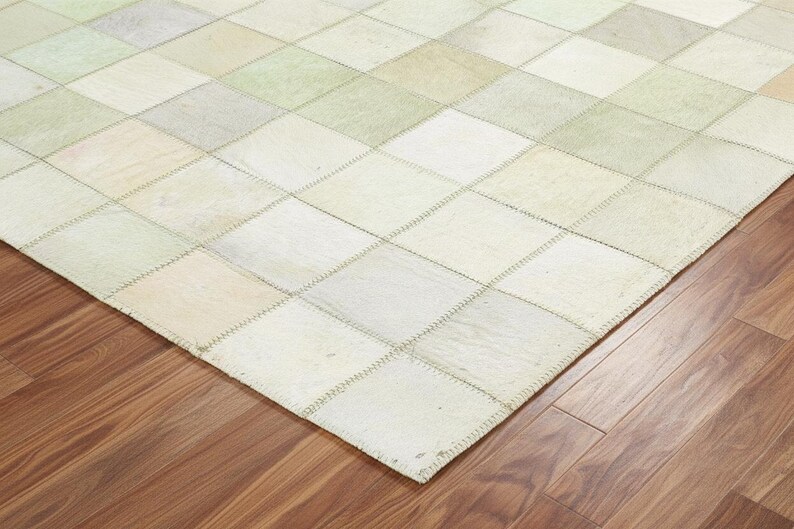 May include: A patchwork rug with a geometric pattern of squares in shades of cream, beige, and pale green. The rug is made of stitched-together pieces and rests on a wooden floor.