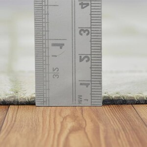 May include: Close-up of a light green rug with a ruler showing measurements in inches. The rug has a stitched border and a grid pattern. The ruler is silver and has black markings. The rug is on a wooden surface.
