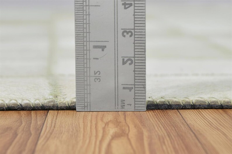May include: Close-up of a light green rug with a ruler showing measurements in inches. The rug has a stitched border and a grid pattern. The ruler is silver and has black markings. The rug is on a wooden surface.