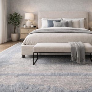 May include: A serene bedroom scene featuring a large bed with a neutral-toned headboard and bedding. A bench with a black metal frame sits at the foot of the bed, and a gray throw blanket is draped over it. A patterned rug in shades of blue and gray covers the floor.