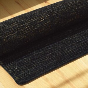 May include: A rolled-up black jute rug with a textured weave. The rug is partially unrolled, revealing its woven construction. The rug is placed on a light-colored wooden floor, with sunlight casting shadows.
