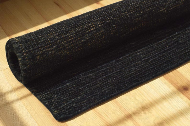 May include: A rolled-up black jute rug with a textured weave. The rug is partially unrolled, revealing its woven construction. The rug is placed on a light-colored wooden floor, with sunlight casting shadows.