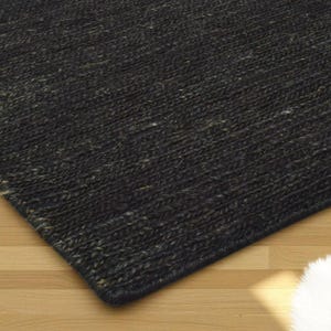 May include: A close-up of a dark gray woven rug with a textured surface. The rug has a rectangular shape and is placed on a light brown wooden floor. The rug's corner is visible.