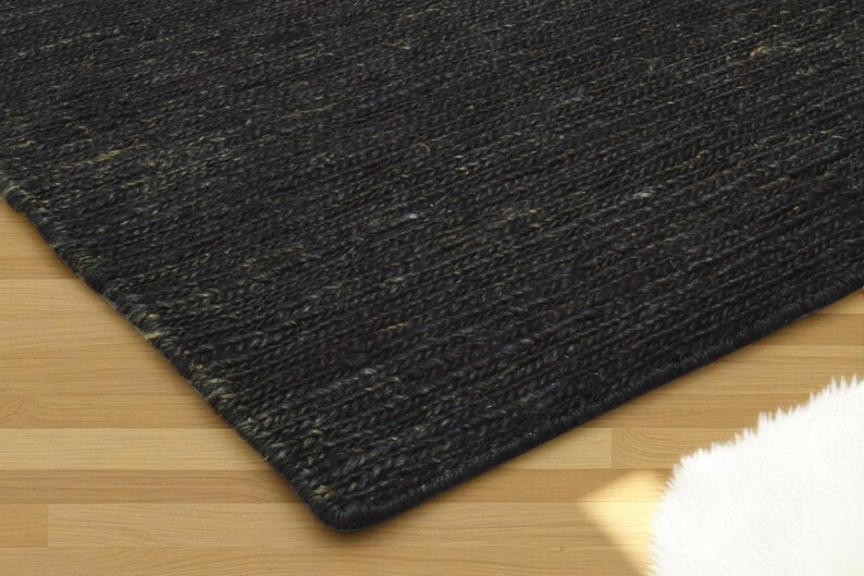 May include: A close-up of a dark gray woven rug with a textured surface. The rug has a rectangular shape and is placed on a light brown wooden floor. The rug's corner is visible.