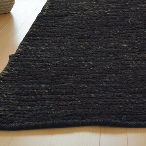 May include: A close-up of a dark gray rug with a braided texture. The rug is made of a thick, woven material and has a slightly uneven surface. The rug is placed on a light-colored wooden floor.