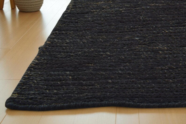 May include: A close-up of a dark gray rug with a braided texture. The rug is made of a thick, woven material and has a slightly uneven surface. The rug is placed on a light-colored wooden floor.