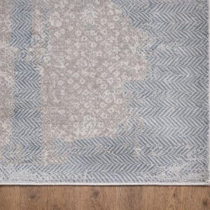 May include: A section of a rectangular area rug with a distressed, vintage appearance. The rug features a combination of gray and light blue hues, with a central pattern and a border with a herringbone design. The rug is placed on a wooden floor.