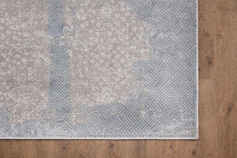 May include: A section of a rectangular area rug with a distressed, vintage appearance. The rug features a combination of gray and light blue hues, with a central pattern and a border with a herringbone design. The rug is placed on a wooden floor.