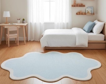 Puede incluir: Una alfombra en forma de nube azul claro con borde blanco sobre un suelo de madera en un dormitorio. La habitación incluye una cama con ropa de cama blanca, un escritorio de madera y una ventana con cortinas blancas. La alfombra añade un toque suave y caprichoso.