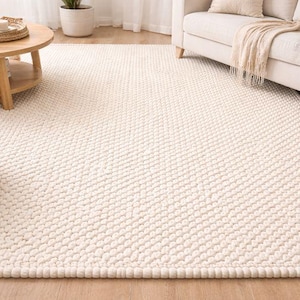 May include: Cream-colored, textured area rug with a dense, looped pile. The rug has a subtle pattern and a thick border. The neutral color and plush texture suggest a cozy and comfortable home decor item. The rug is placed on a wooden floor.