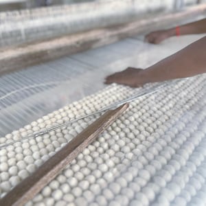 May include: A close-up of a weaving loom in operation. The loom features white threads and a wooden shuttle. The hands of a person are visible, working with the threads. The woven material appears to be a soft, white fabric.
