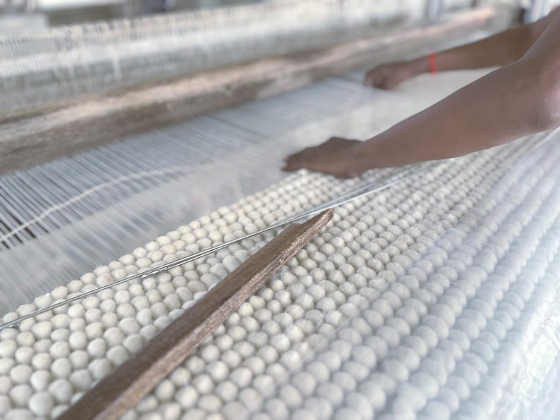 May include: A close-up of a weaving loom in operation. The loom features white threads and a wooden shuttle. The hands of a person are visible, working with the threads. The woven material appears to be a soft, white fabric.