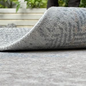 May include: A rolled-up area rug with a gray and blue patterned design. The rug's edge is curled up, revealing the textured underside. The rug is made of a soft, plush material, suitable for indoor use.