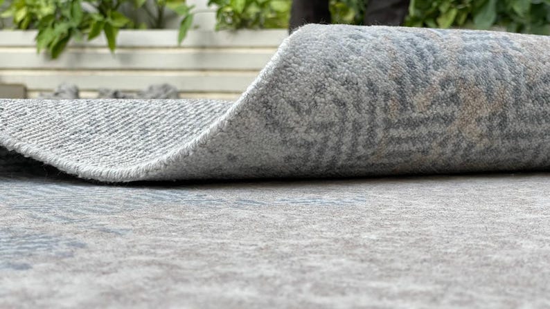 May include: A rolled-up area rug with a gray and blue patterned design. The rug's edge is curled up, revealing the textured underside. The rug is made of a soft, plush material, suitable for indoor use.