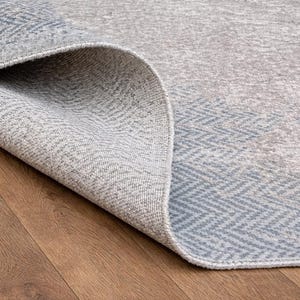 May include: Close-up of a rolled-up area rug with a gray and blue geometric pattern. The rug's edge reveals a textured weave, and the background shows a wooden floor.