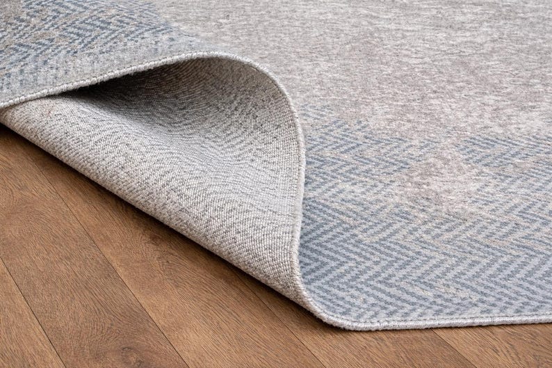 May include: Close-up of a rolled-up area rug with a gray and blue geometric pattern. The rug's edge reveals a textured weave, and the background shows a wooden floor.