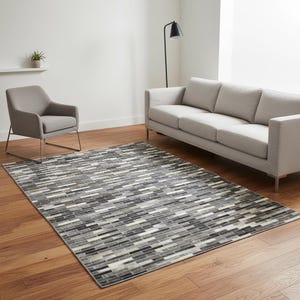 Pode incluir: Um tapete retangular com um padrão geométrico de blocos retangulares cinza, preto e branco. O tapete está colocado sobre um piso de madeira, com um sofá cinza e uma poltrona ao fundo.