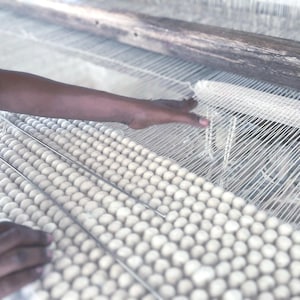 May include: Close-up of a hand weaving on a loom. The loom features white threads and a wooden frame. The woven material appears to be a light-colored, textured fabric. The image highlights the process of textile creation.