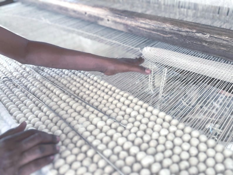 May include: Close-up of a hand weaving on a loom. The loom features white threads and a wooden frame. The woven material appears to be a light-colored, textured fabric. The image highlights the process of textile creation.