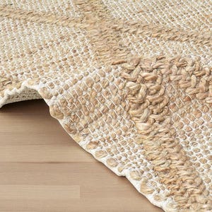 May include: Close-up of a natural fiber rug with a woven design. The rug features a combination of beige and off-white tones, with a textured, braided border. The rug is partially rolled up, revealing the underside.