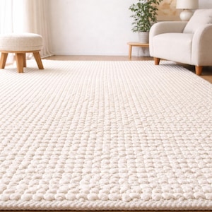 May include: Cream-colored, textured area rug with a repeating pattern of raised, rounded shapes. The rug fills the foreground, with a glimpse of a light-filled room in the background. A wooden floor is visible.