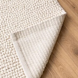 May include: Cream-colored, textured rug with a raised, dotted surface. The corner of the rug is lifted, revealing a woven backing. The rug is placed on a wooden floor, showcasing its soft texture and design. This rug is suitable for home decor.